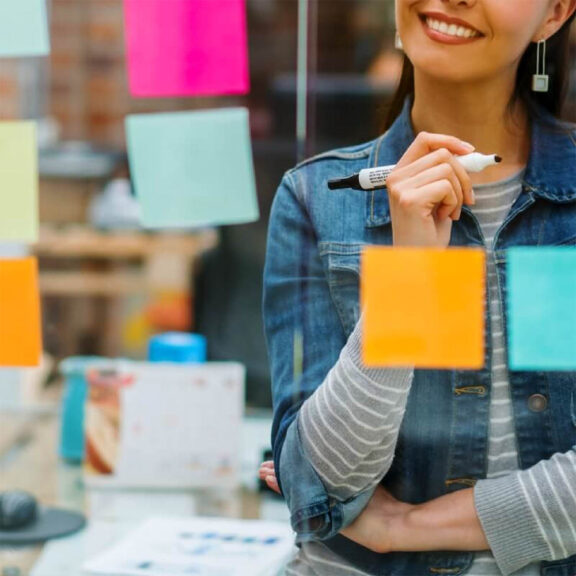 Woman smiling and holding dry erase marker as she looks at glass board with various colors of sticky notes
