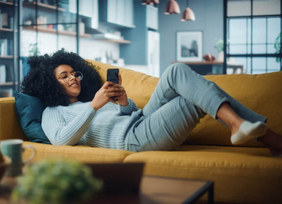 woman with glasses laying down on a light brown couch looking at her phone
