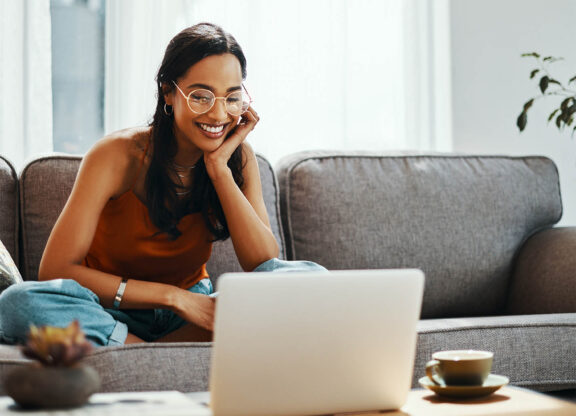 woman with glasses sitting on a grey couch smiling and looking at a laptop screen
