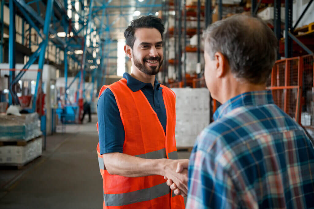 Image of a male manager wearing a plaid shirt shaking the hands of his male warehouse employee who is wearing an orange safety vest