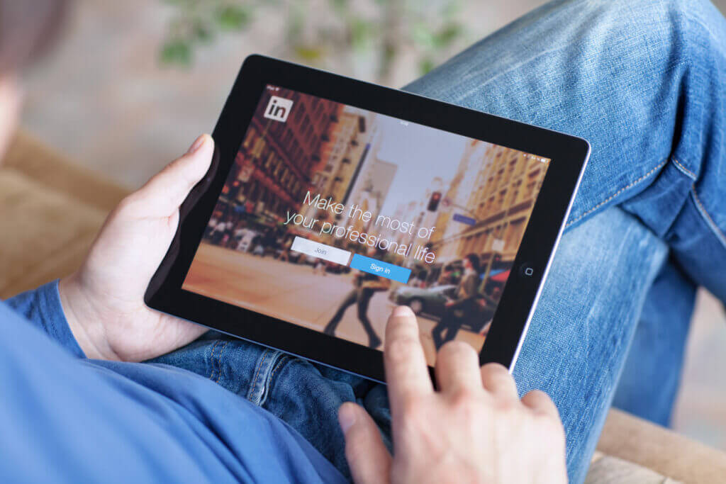 Image of a person holding an iPad with their legs crossed clicking on a LinkedIn social media page