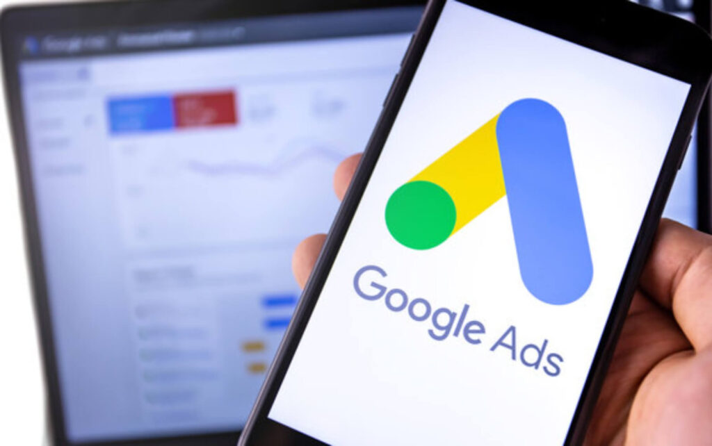 Photo of a person’s hand holding a smartphone with the Google Ads logo displayed on the screen with a computer in the background with the actual Google Ads platform displayed on the screen