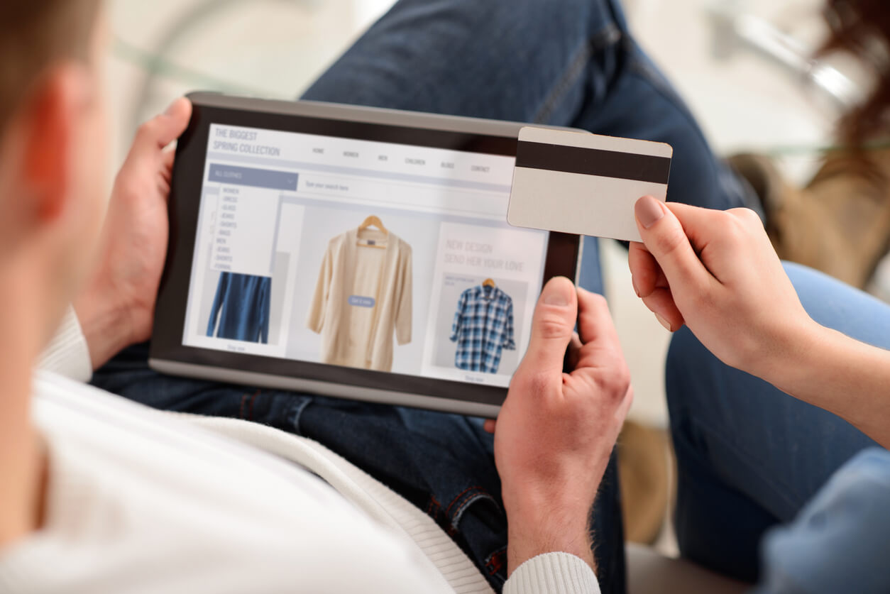 Photo of a tablet with an e-commerce clothing website open as someone scrolls with their finger and holds a credit card